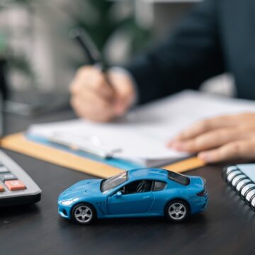 Concept Image Of The Car Buying Process Showing A Person Filling Out Paperwork With A Blue Model Car On The Desk