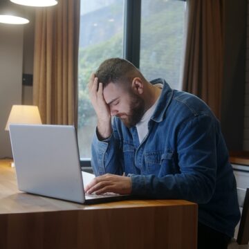 A Stressed, Bearded, Man Sitting At The Kitchen Table Using A Laptop And Holding His Head In His Hand