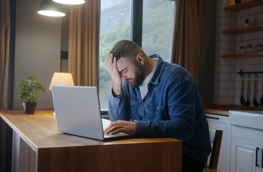 A Stressed, Bearded, Man Sitting At The Kitchen Table Using A Laptop And Holding His Head In His Hand A Stressed, Bearded, Man Sitting At The Kitchen Table Using A Laptop And Holding His Head In His Hand