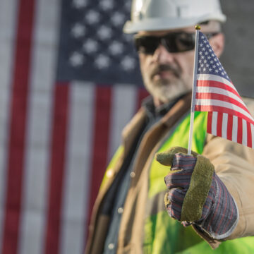 A Construction Worker Wearing A Hard Hat, Holding A Small American Flag, And Standing In Front Of A Large American Flag