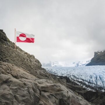 A Photo Of The Greenland National Flag Flying Above A Rocky Arctic Glacier And Mountains
