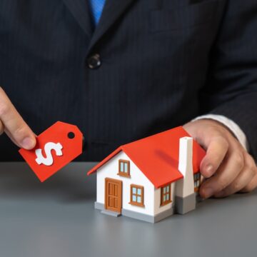 A Person In A Business Suit Pictured With A Small Model House And A Price Tag, Symbolizing The Process Of Valuing Property