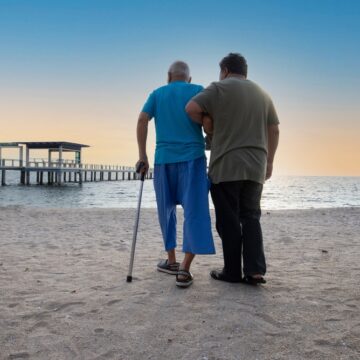 A Younger Person Assisting An Older Person, Who Is Using A Walking Stick, On A Sandy Beach At Sunset