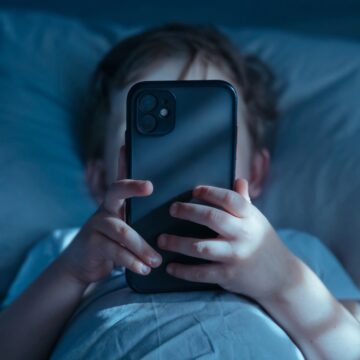 A Photo Of A Young Child Using A Smartphone In Bed At Night, Illuminated By The Screen's Blue Light