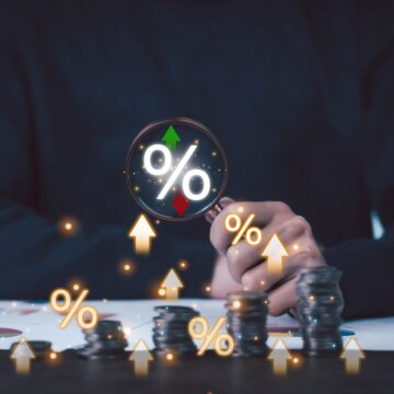 a businessman using a magnifying glass to highlight a percentage symbol over stacks of coins, representing the scrutiny or monitoring of financial growth and market trends