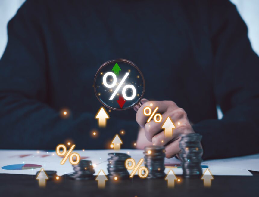 a businessman using a magnifying glass to highlight a percentage symbol over stacks of coins, representing the scrutiny or monitoring of financial growth and market trends