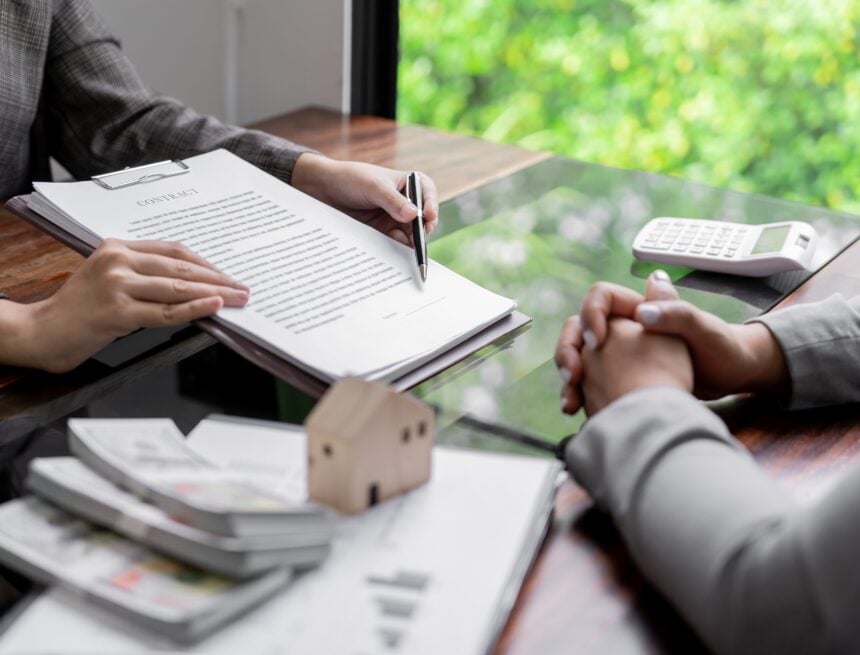 Image of people signing a contract with real estate icons such as a small house model, paperwork, and stacks of currency.