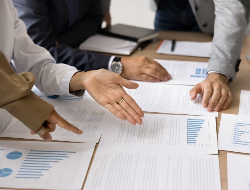 A close up of a group of businesspeople looking over documents, bar charts, and pie graphs spread across a table.