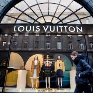 A Woman Walks In Front Of The Luxury Boutique Louis Vuitton Brand Store And Window Display With Mannequins.
