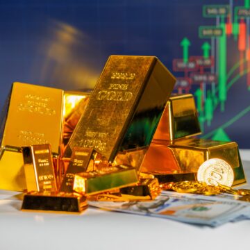 The image shows safe-haven asset gold bars, bouillon, and coins, atop currency with upward graphs in the background.