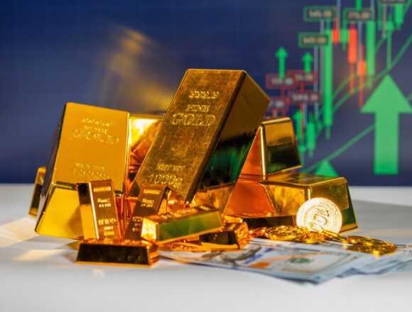 The image shows safe-haven asset gold bars, bouillon, and coins, atop currency with upward graphs in the background.