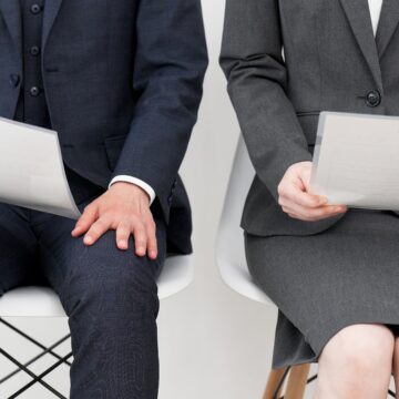 An image of a man and a woman, dressed in business attire and sitting and waiting in chairs while holding papers presumed to be resumes.