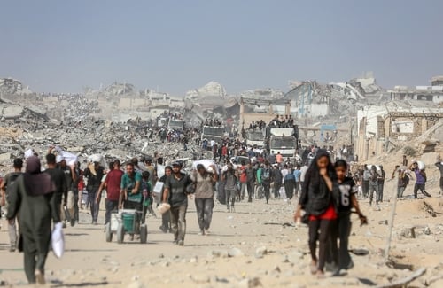 Displaced Palestinian civilians carry flour and food aid on a debris-strewn road amidst extensively damaged buildings in the Gaza Strip.