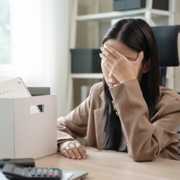A distressed woman, experiencing job loss, with her hand over her face and a box containing personal belongings, such as a calendar and office supplies, visible on the desk.