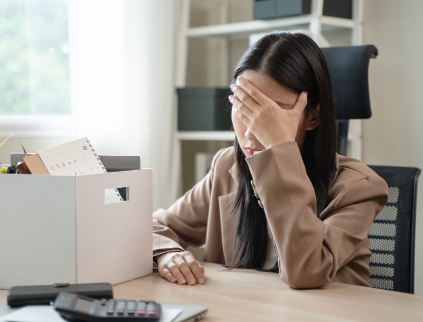 A distressed woman, experiencing job loss, with her hand over her face and a box containing personal belongings, such as a calendar and office supplies, visible on the desk.