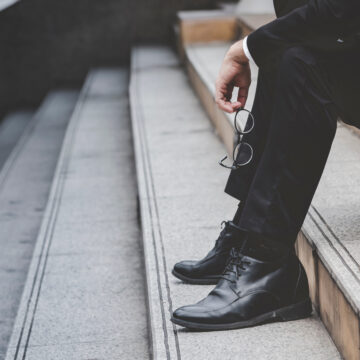 The feet and the legs of a businessman sitting dejectedly on a set of stairs, likely experiencing stress, failure, or unemployment.