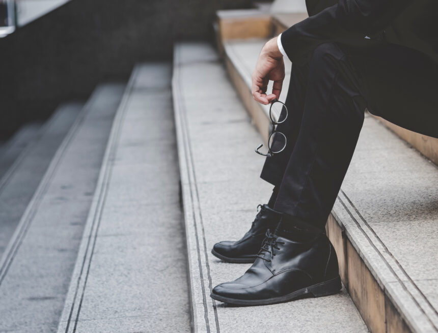 The feet and the legs of a businessman sitting dejectedly on a set of stairs, likely experiencing stress, failure, or unemployment.