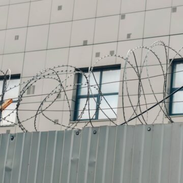 Metal,Fence,With,Barbed,Wire,Around,Building. LOCKED DOWN. ROUNDED UP. WAREHOUSED. THE RISE OF AMERICA’S CONCENTRATION CAMPS