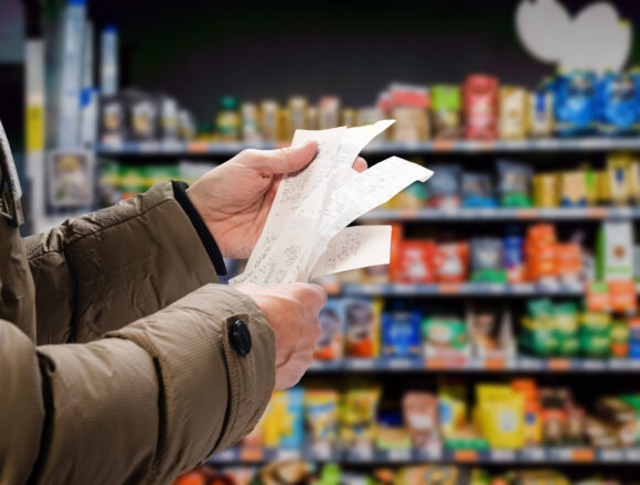 A shopper reviewing long receipts in front of stocked supermarket shelves, representing the increasing cost of living.