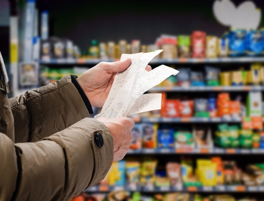 A shopper reviewing long receipts in front of stocked supermarket shelves, representing the increasing cost of living. A shopper reviewing long receipts in front of stocked supermarket shelves, representing the increasing cost of living.