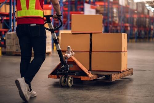 A factory worker pushing a loaded hand pallet truck in a warehouse or distribution center.