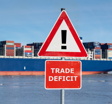 A red warning sign with the text "Trade Deficit" stands on shore with a loaded cargo ship at sea in the background. A red warning sign with the text "Trade Deficit" stands on shore with a loaded cargo ship at sea in the background.