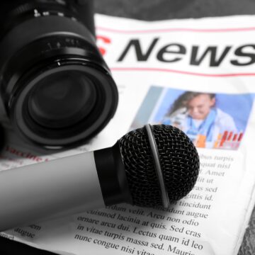 An image of the essential tools used in journalism, featuring a microphone resting on a newspaper alongside a camera.