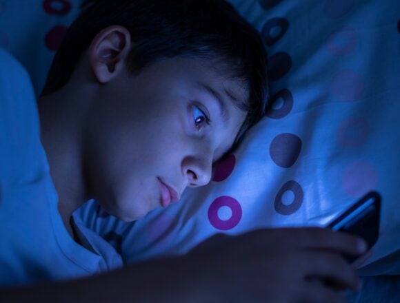 A boy looking at a mobile phone in bed at night, illuminated by the screen's blue light.