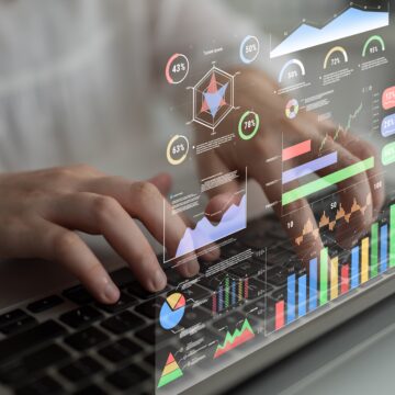 A futuristic heads-up data analytics display superimposed over a person typing on a laptop showing financial metrics, stock ticker, colorful charts, graphs, and percentage indicators for monitoring business performance.