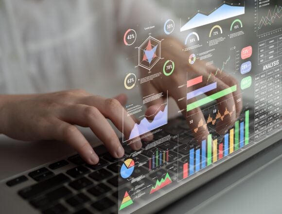 A futuristic heads-up data analytics display superimposed over a person typing on a laptop showing financial metrics, stock ticker, colorful charts, graphs, and percentage indicators for monitoring business performance.