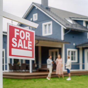 A young couple visiting a potential new home with a "For Sale" sign in the front yard.