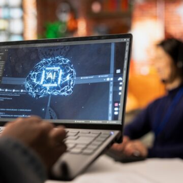 Image Shows A Person Working On A Laptop That Is Running Software Related To Artificial Intelligence (AI), As The Screen Shows A Graphic Of A Brain With "AI" In The Center And Lines Of Code Or Data.