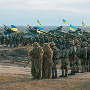 Photo Of Ukrainian Armed Forces Soldiers And Military Equipment During A Training Exercise, Some Flying Ukrainian Flags, Lined Up In A Field Setting, Wearing Military Camouflage And Specialized, Full-body Ghillie Suits, Used For Advanced Camouflage By Snipers And Reconnaissance Personnel.