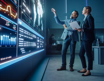 A data analytics center with two professionals in business attire collaborating while analyzing complex data displayed on a large video wall.