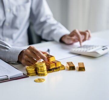 Businessman counts gold bars and coins and analyzes cost with adding machine.