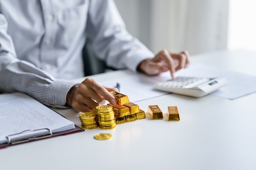 Businessman counts gold bars and coins and analyzes cost with adding machine.