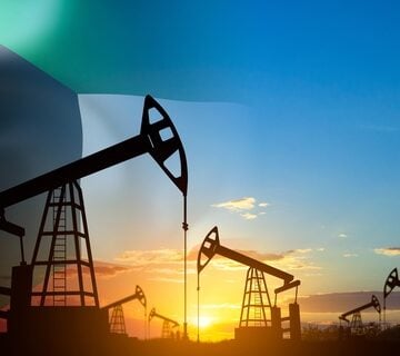 A field of oil pump jacks, silhouetted against a sunrise or sunset, with the flag of Kuwait overlaid on the left side.
