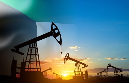 A field of oil pump jacks, silhouetted against a sunrise or sunset, with the flag of Kuwait overlaid on the left side.