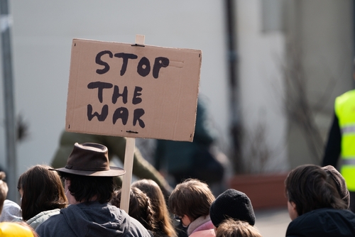 A protest sign reads "STOP THE WAR" in hand-lettered black text on cardboard, being held up by a person in a crowd during a public demonstration.