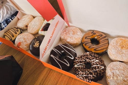A box of Dunkin' Donuts containing a mix of classic and flavored varieties, including powdered sugar, chocolate-frosted, and sprinkle-topped doughnuts.
