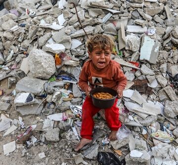 This image shows a young Palestinian boy crying while sitting amidst the rubble of destroyed buildings in Gaza.