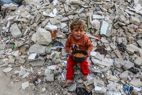 This image shows a young Palestinian boy crying while sitting amidst the rubble of destroyed buildings in Gaza.