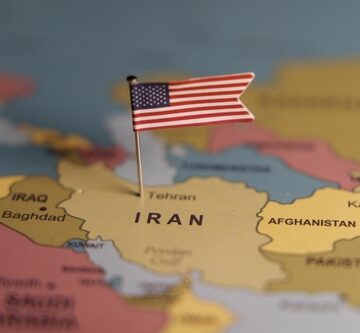 An image of a map of Iran with a U.S. flag pinned into it symbolizing political conflict.
