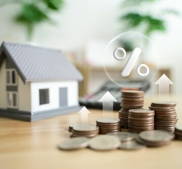 Rising mortgage rate concept with a miniature house, stacks of coins, and a percentage sign next to an upward arrow.