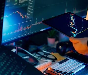 A trader performing financial analysis of stock or cryptocurrency market charts on a computer monitor and a tablet.