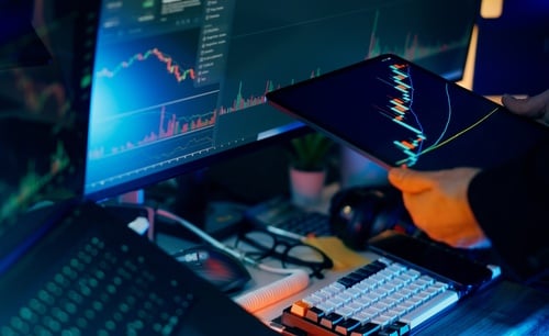 A trader performing financial analysis of stock or cryptocurrency market charts on a computer monitor and a tablet.