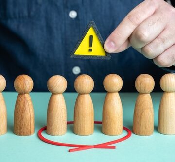 Wooden Figures in a row, with a red circle around some and a yellow triangle with an exclamation point being held above, identifying this specific group as currently at risk.