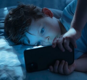 A young boy, lying in bed in a dark room, is bathed in the blue light emitted from the smartphone screen he's using.