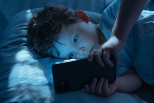 A young boy, lying in bed in a dark room, is bathed in the blue light emitted from the smartphone screen he's using.