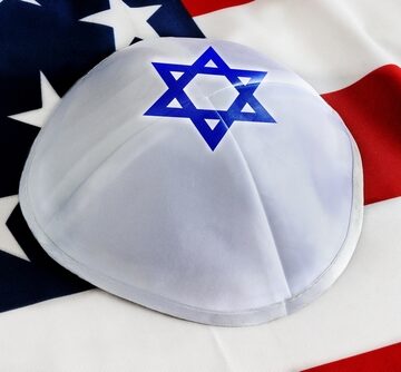 A white satin yarmulke, featuring a blue Star of David, with an American flag in the background.